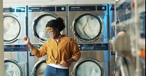 Beautiful African American cheerful woman in stylish outfit having fun and dancing in laundry service room while machines washing on background. Girl resting while machines working in washhouse.