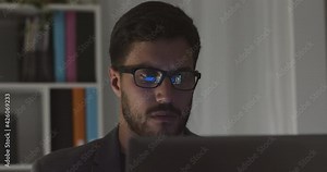 Young caucasian handsome businessman in eye glasses late at night scrolling in front of computer. Financial animations reflected in glasses. Male broker following rates and bets.