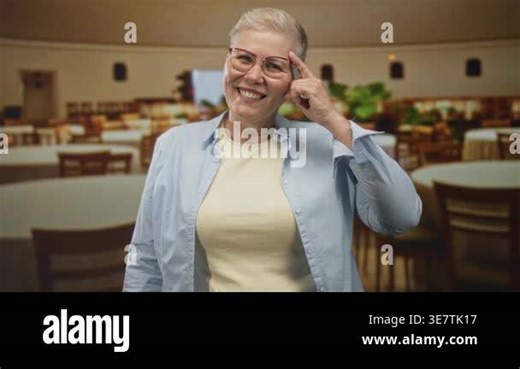 Woman wearing glasses points finger to temple while smiling in restaurant dining room with round tables and chairs; friendly joy Stock Video Footage - Alamy