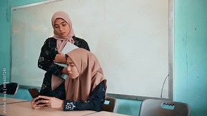 Two beautiful asian muslim women smiling happily relaxing using laptop computer, working, discussing project plans and exchanging ideas in the classroom.
