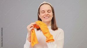 Positive housekeeping. Cheerful female housewife wearing hair band and white sweater taking off orange rubber gloves posing isolated over gray background being happy to finish cleaning her house