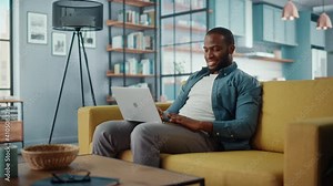 Handsome Black African American Man Working on Laptop Computer while Sitting on a Sofa in Cozy Living Room. Freelancer Working From Home. Browsing Internet, Using Social Networks, Having Fun in Flat.