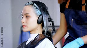 Portrait of young Asian female sitting in modern parlour while getting hair colouring done wearing face mask for protection with female hairdresser colouring hair