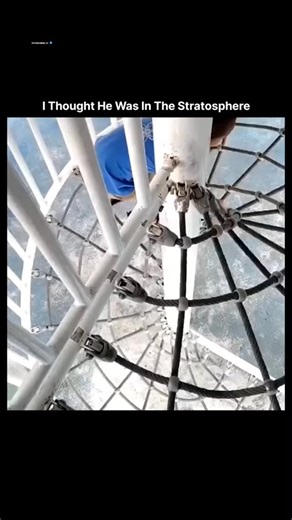 science 🔭 on Instagram: "This Instagram Reel depicts a young Asian boy, approximately 4-5 years old, dressed in a blue graphic T-shirt and sandals, methodically climbing a tall spiral staircase constructed from white cargo netting. The structure appears suspended over a blue-tinted surface, possibly a pool or padded area, creating an optical illusion of extreme height against a bright outdoor setting with red flooring and white railings. The footage captures his determined hand-over-hand ascent