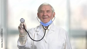 Front view old man doctor with sthetoscope smiling. Show stethoscope for treatment patient at hospital. Blurreed indoor background.