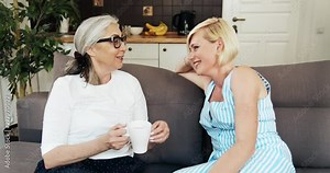 Joyful middle aged lady visiting retired mother, both sitting on comfortable couch and having pleasant chat and drinking coffee