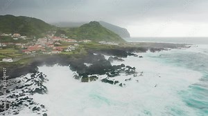 Flores Island, Azores, Portugal. Aerial view of pebbled beach washed with foamy waves of the Atlantic Ocean. Remote, charming settlement beneath rocky green cliffs. High quality 4k footage