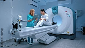 Doctor holding folder and making notes of patient symptoms. Patient lying at CT scanner. Modern equipped cabinet in private clinic. Doctor talking to patient before procedure. MRI examination.