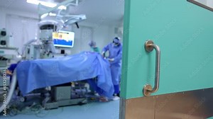 Ajar doors to a modern surgery room. Surgeon works at the head of the patient. Blurred backdrop.