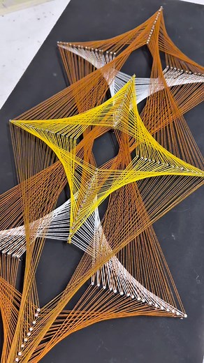 Pin art with strings 📌 This piece is a vibrant exploration of symmetry and dimensionality using the medium of string and pins. The artwork is composed of meticulously arranged threads—primarily in yellow, orange, and white tones—woven across strategically placed nails on a black canvas. The result is a captivating diamond-shaped pattern that gives the illusion of depth and motion. #StringArt #GeometricDesign #ThreadArt #OpticalIllusionArt #ContemporaryArt #MandalaInspired #SymmetryInArt #Handma