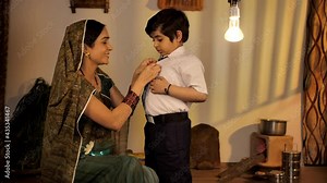 Village woman helping her son to get ready for school - parent-child relationship and care. Medium shot of a rural mother in traditional Saree and son in school uniform - Indian village. Child educ...