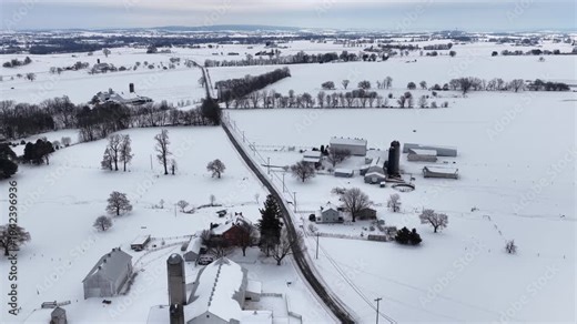Aerial winter landscape of rural Pennsylvania farmland with barns, silos and country road crossing snow-covered agricultural fields. Descend wide shot. Peaceful calm suburbia of town.