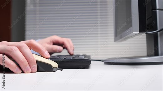 Person's hands operating a computer mouse and typing on a keyboard, with a monitor visible on a clean white desk, representing office work and technology
