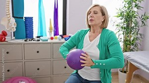 Mature woman exercises with a purple ball in a brightly lit physical therapy clinic, promoting wellness and active aging.