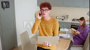 Young couple working remotely from home. Redhead man in glasses making business phone call holding open laptop, his wife browsing through notepad in background