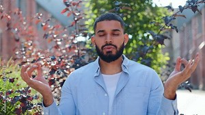 Front view of male yoga practitioner clasping fingers in mudra position, closing eyes, breathing out and doing meditation exercise. Casually dressed arabian man taking look at camera and smiling.
