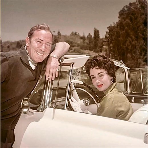 "Elizabeth Taylor and her husband, Michael Wilding, are photographed sharing a lighthearted conversation from the window of her car outside their Los Angeles home in 1955. At the time, Taylor was one of Hollywood’s most beloved actresses, while Wilding, a distinguished British actor, had recently moved to California to be with her. Married in 1952, the couple was frequently seen around Beverly Hills, often drawing admiration for their grace and poise. This candid moment, captured just before Tay