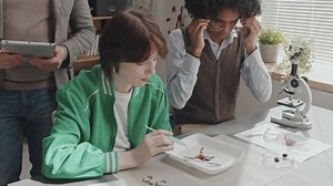 Tilting down of bearded male teacher holding tablet, standing in classroom, watching two high school classmates sitting at desk, using microscope and dissecting frog
