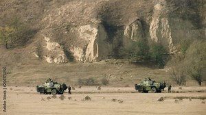 two military combat vehicles in the field under the sand hills with an army around them