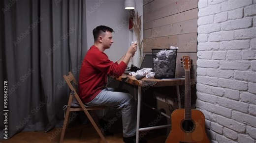 Man at desk crumpling paper in frustration, surrounded by discarded notes and a guitar, struggling with writers block and creative exhaustion while trying to compose music or lyrics
