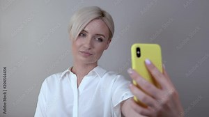 Closeup of cute shorthaired blonde girl poses for selfies. Woman makes a selfie using a phone in a yellow case.