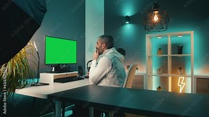Bearded man in denim vest clicks and scrolls on large computer monitor with chroma key in vibrant home office. He uses an ergonomic keyboard and mouse to work efficiently and comfortably.