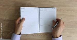 Man flips pages of diary calendar holding pen in hand. Male manager ready to write working schedule sitting at wooden table in office