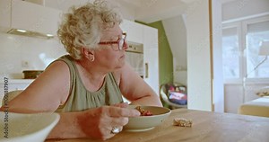 An elderly woman sits alone at her kitchen table eating a healthy breakfast of granola and berries.