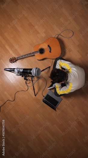 Vertical video creator setting up a digital audio workstation, connecting a microphone and acoustic guitar to a laptop on a wood floor, preparing to record new music or produce original songs from hom