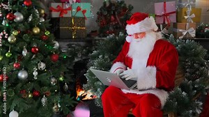 Smart Santa uses laptop typing on keyboard in his cottage near Christmas tree and cozy fireplace.