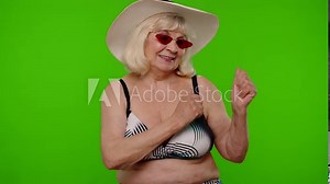 Senior pensioner woman tourist in swimsuit bra, red sunglasses and hat dancing, celebrating, smiling, posing on chroma key background. Portrait of exited old granny traveler model on summer vacations