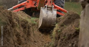 A low angle view of a small backhoe digging a drainage ditch. With audio.