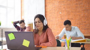 young attractive woman student sitting in front of computer in big white headphones. A woman in an office or a classroom. in the background people are sitting. Online communication. A girl drinks Stock Video