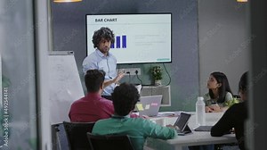 Young Indian businessman corporate leader coach speaker give flip chart workshop presentation explaining strategy teaching multi ethnic staff consult diverse team at office meeting training.