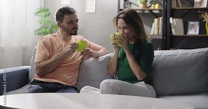 Cheerful couple talking drinking tea or coffee together in the living room. Loving wife and husband share news, enjoy communication. Romantic relationship, intimate connection