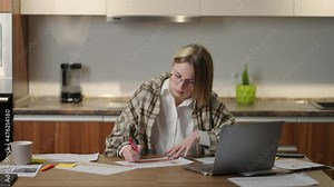 A woman with glasses works remotely from home sitting at a table with a laptop and a felt-tip pen marks the data on the graph. Distance learning student at home to perform an assignment in economics