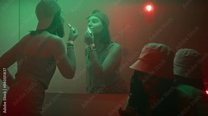 Handheld shot of friendly Black girl with cellphone greeting young Caucasian lady who entering smoky restroom to apply lipstick while hanging out in nightclub in y2k outfits
