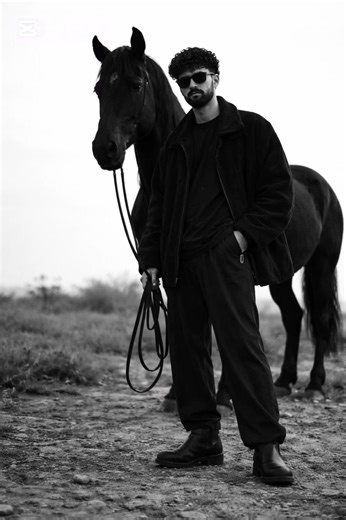 #sargodha❤️ @Ibs. Leader #pubgmobile #ai #prompt high-fashion black-and-white editorial photograph of a person sitting low on a minimalist metal stool, legs spread forward in a relaxed, grounded pose, feet planted on uneven outdoor terrain. One hand loosely resting between the knees holding thin leather reins, shoulders slightly slouched, head tilted downward in a calm, introspective expression. The subject wears dark oversized layered clothing, wide-leg trousers, leather boots, and dark sunglas