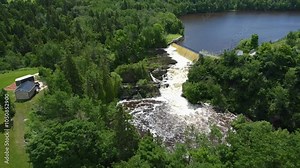 Aerial view looking down towards the waterfall of an old abandoned hydroelectric power plant dam from which a good flow of oxidized yellowish water flows through a lush green forest. Riviere-du-loup.