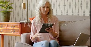 Senior woman checking laptop on sofa grabbing papers and scrolling tablet while reviewing documents