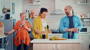 Young man making proposal to his girlfriend in front parents while they talking in the kitchen at home on family day. She putting ring on her finger, kissing and hugging him, mother take pictures