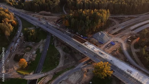 Drone follows diagonal bridge axis as traffic moves and crews work on new concrete decking, cloverleaf ramps, and trailers near suburban forest and gravel piles.