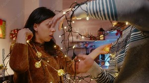 Couple laughing and getting tangled in string of christmas lights. Man and woman trying to untangle knot of garland with twinkle lights and illuminated bulbs. Holiday decorations