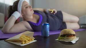 Lazy lady in sportswear lying on yoga mat eating junk food instead of training