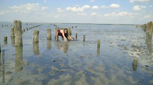 Slender blonde with long hair in a red dress in mud poses in the water against a background of clouds holding on to wooden poles for salt pools and splashing water and picking up dirt from the estuary