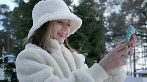 Young woman in white hat and fur coat using mobile phone at winter street. Smiling girl in white fur coat and hat browsing smartphone on fit tree background at winter