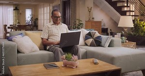 Elderly Indian Man Using Laptop at Home: Using Online Platforms to Explore His Interests, Delving into History, Arts, and Literature. Cultivating Wisdom and Sharing Stories On Social Media. Wide Shot