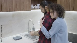 Glad wife hugging husband, washing dishes in the kitchen, family care, help
