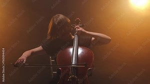 Close up of skilled female hands holding bow and playing classic music on cello. Woman performing musical show on stage and creating breathtaking melody while sitting on chair in bright spotlights.