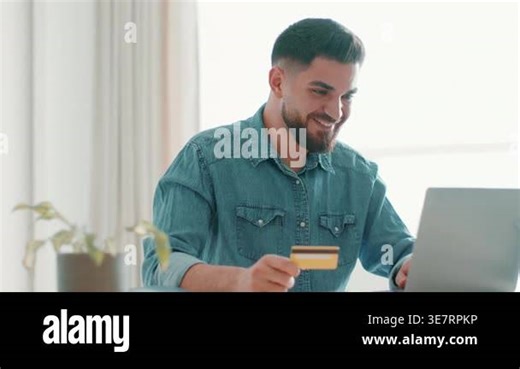 A man with dark skin sits at a table using a laptop. He holds a credit card in his hand and smiles while looking at the computer screen. A plant is on the table, and sunlight comes through the window Stock Video Footage - Alamy
